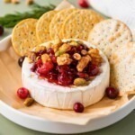 A round block of brie cheese, topped with nuts and cranberries, surrounded by crackers.