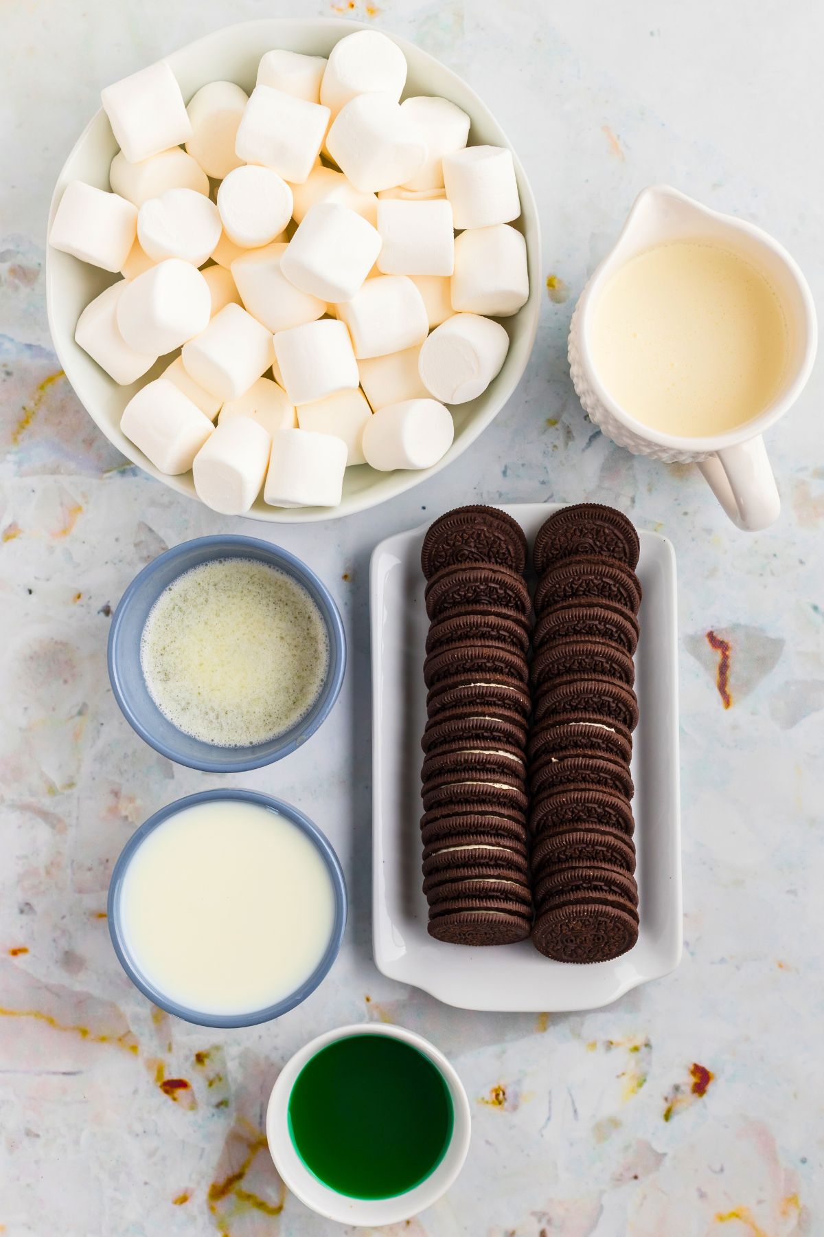 Chocolate cookies, marshmallows, and other ingredients measured into small containers on a marble table. 