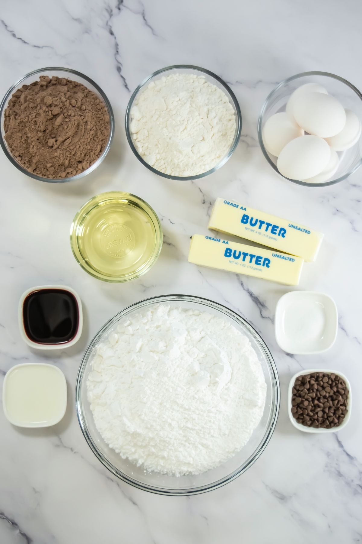 Ingredients needed to make cupcakes and frosting measured in small glass bowls on a marble table. 