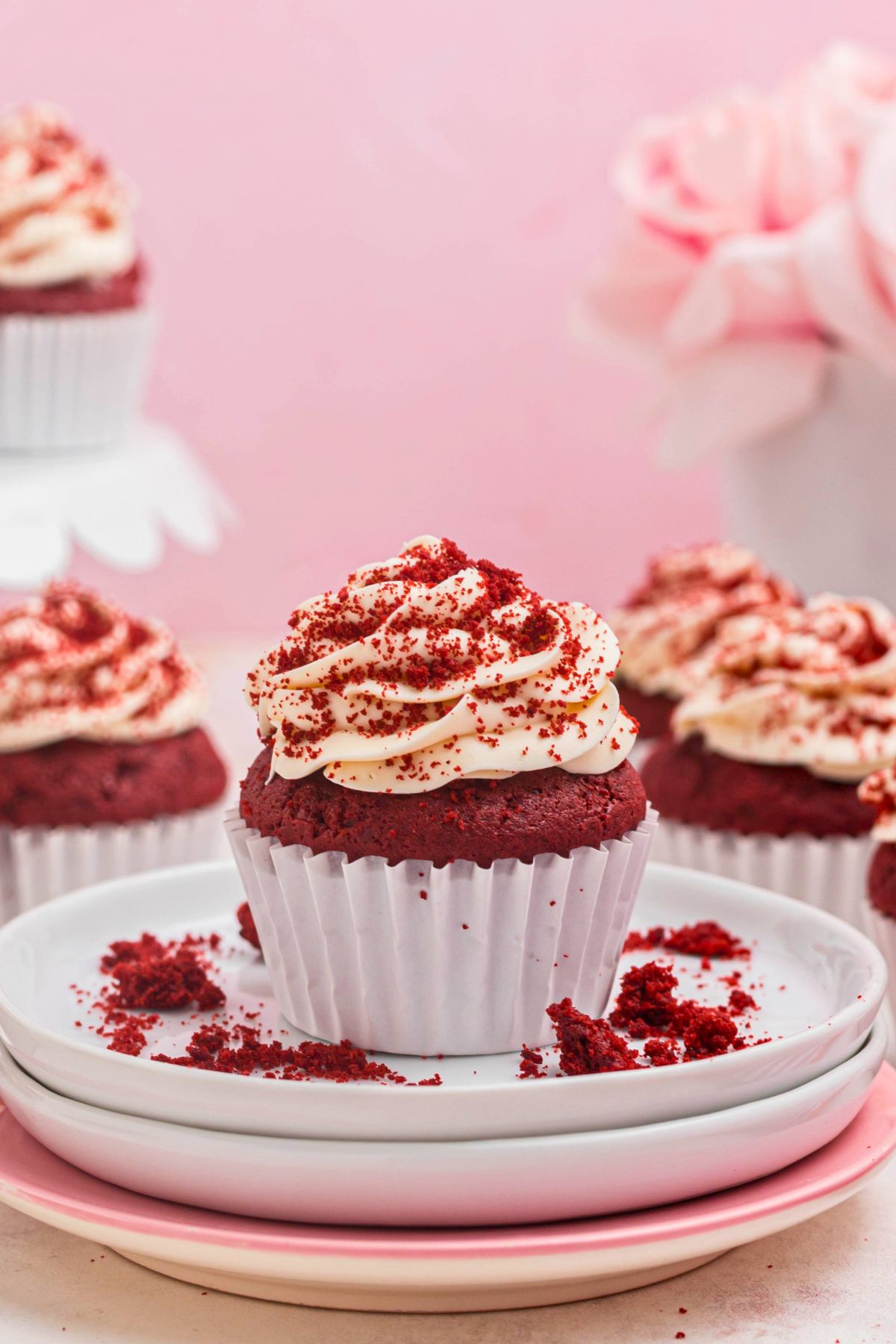 Bright red velvet cupcakes on white plate with cupcakes on a stand with a pink wall behind them. 