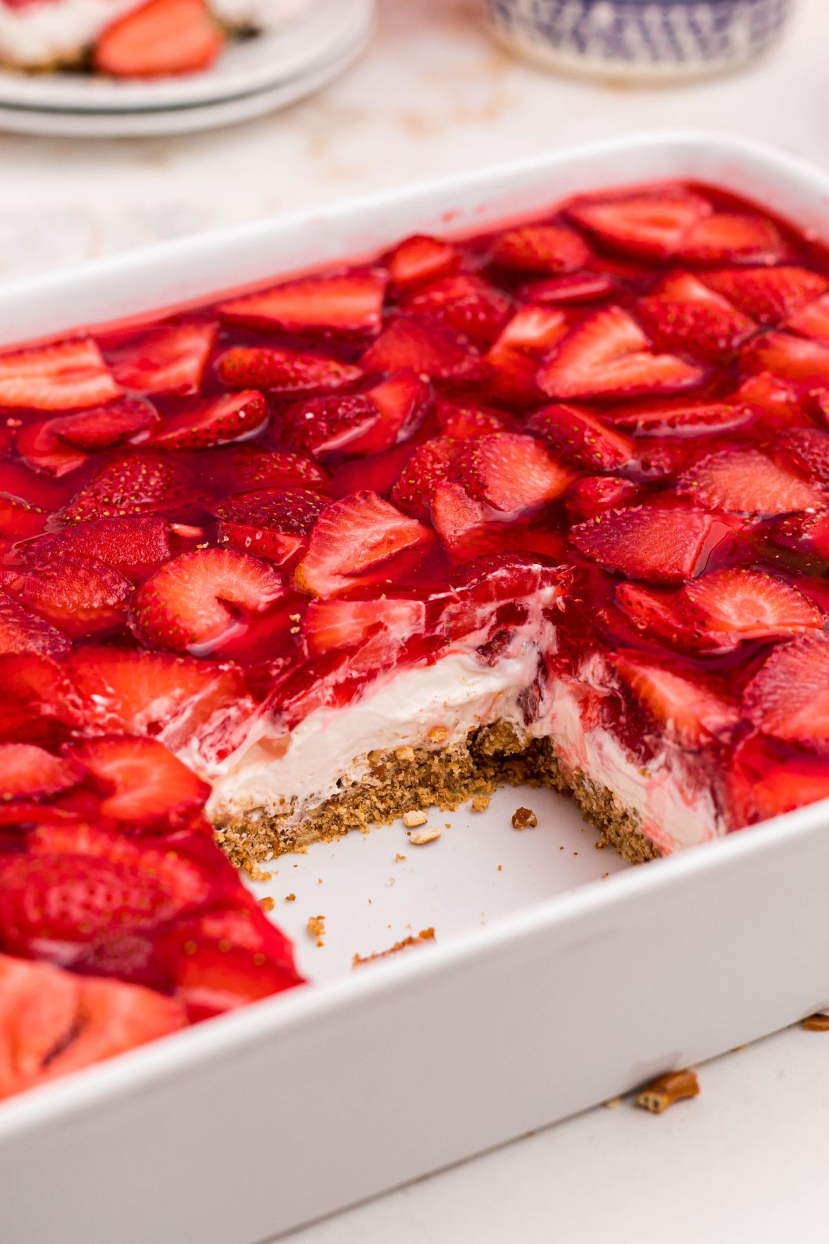 Layers of pretzel crust, whipped filling, and strawberries in J-ello, in a white baking dish with a slice missing. 