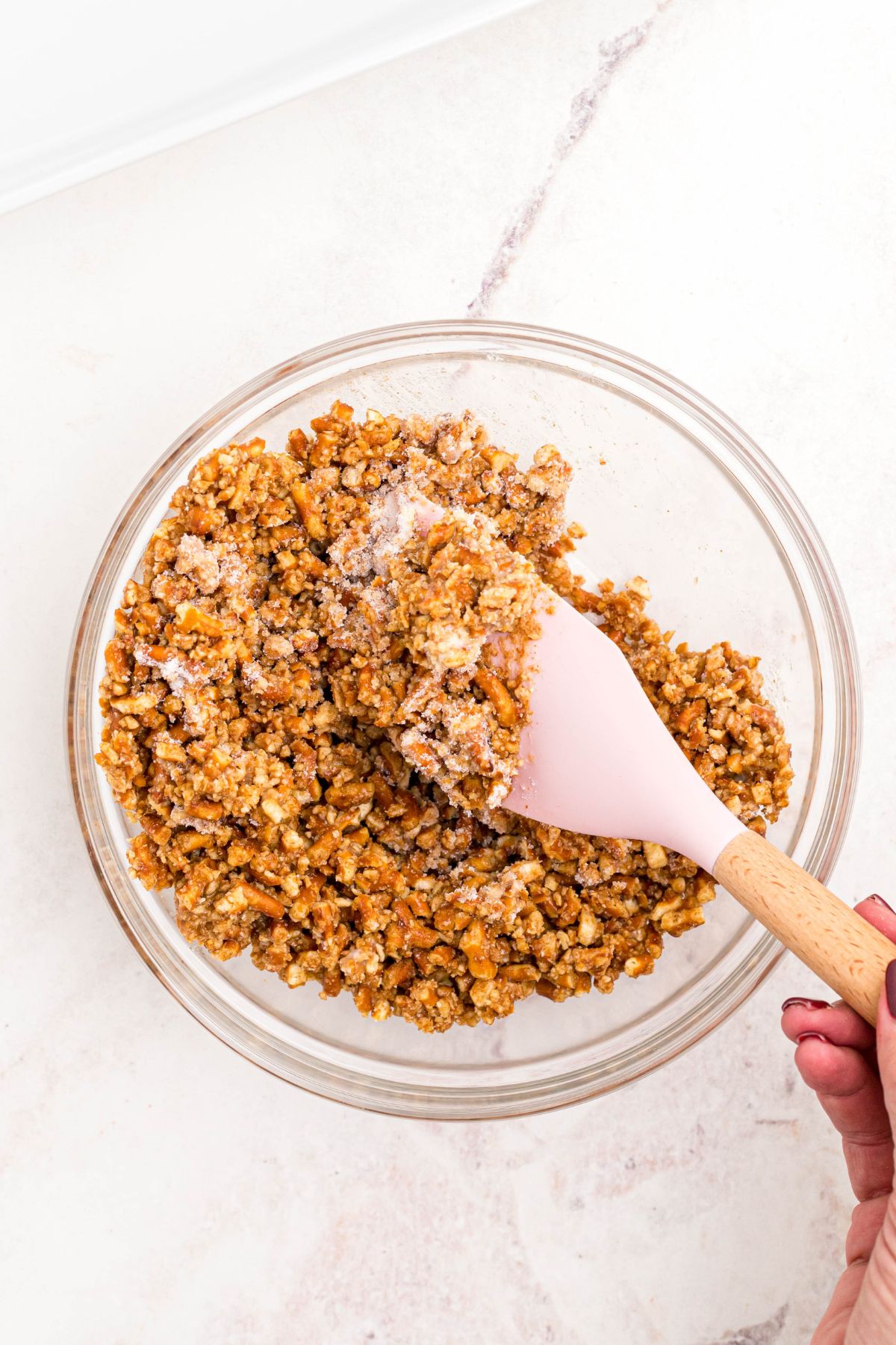 Crushed pretzels being mixed with melted butter and sugar, in a clear glass bowl.
