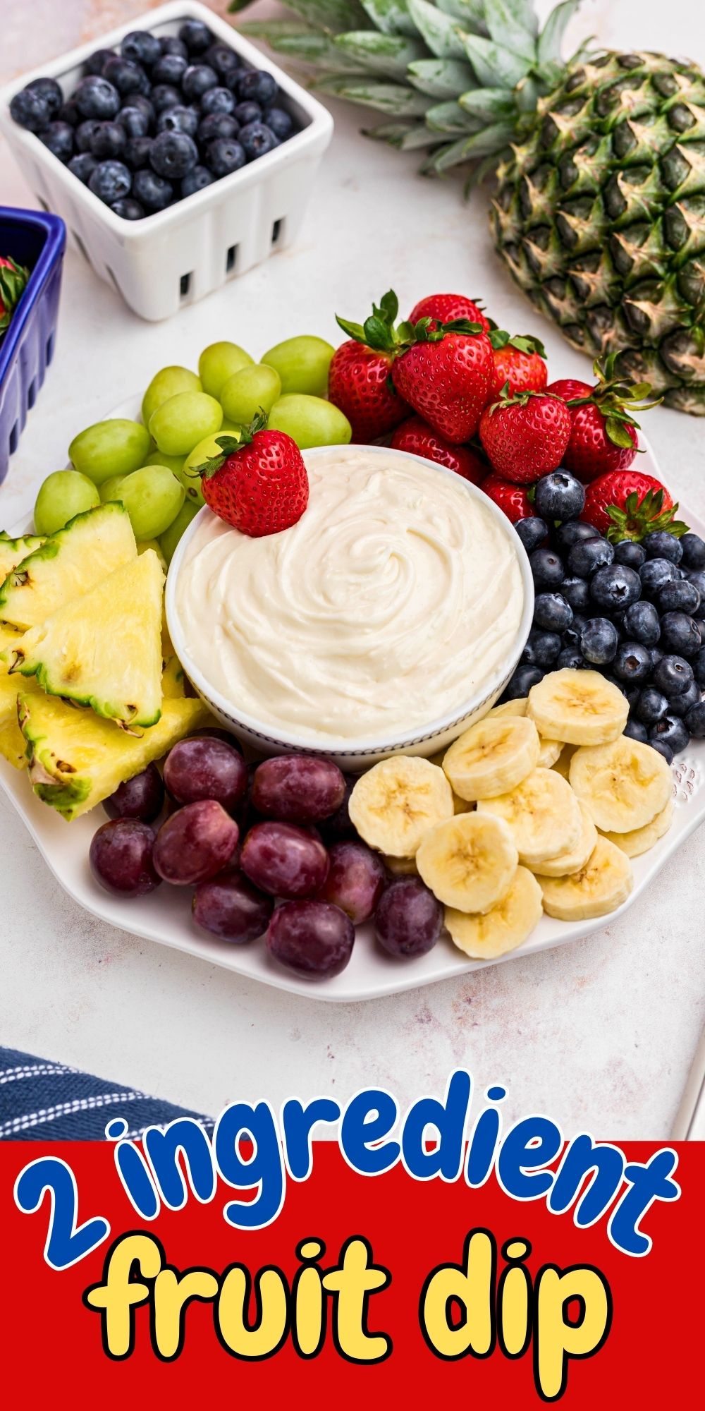 Bright berries, grapes, bananas, and pineapple, around a small bowl with fruit dip in the center of a large white platter.