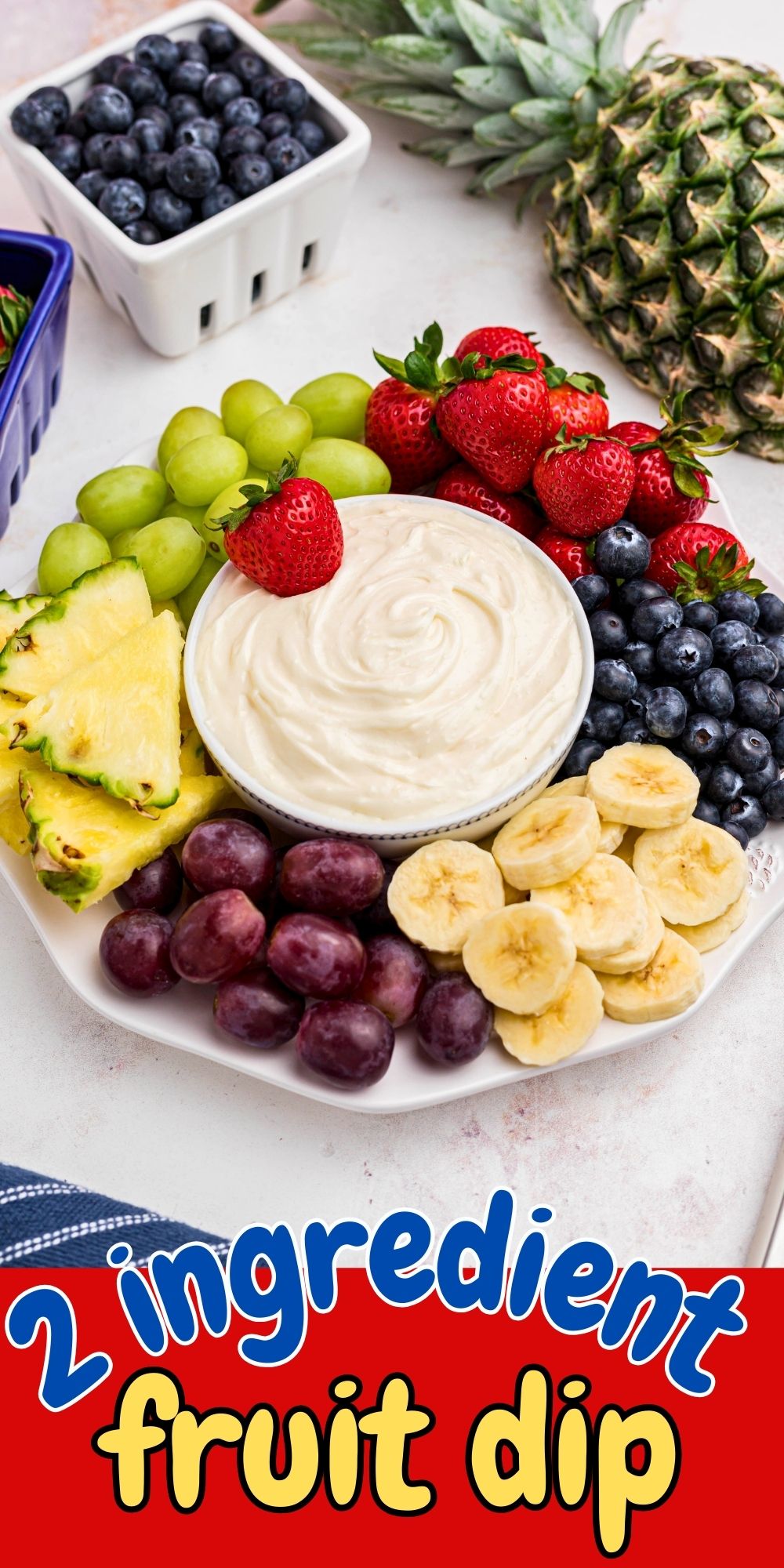 Bright berries, grapes, bananas, and pineapple, around a small bowl with fruit dip in the center of a large white platter.