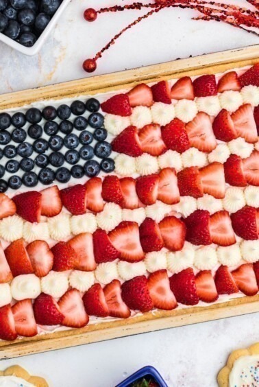 Rectangle cookie in cookie pan, topped with frosting, blueberries and strawberries, on a marble table.