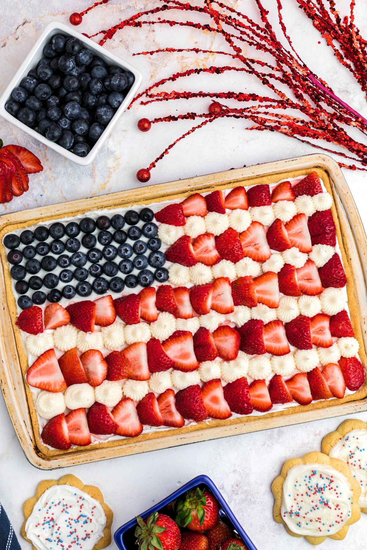 Rectangle cookie in cookie pan, topped with frosting, blueberries and strawberries, on a marble table. 