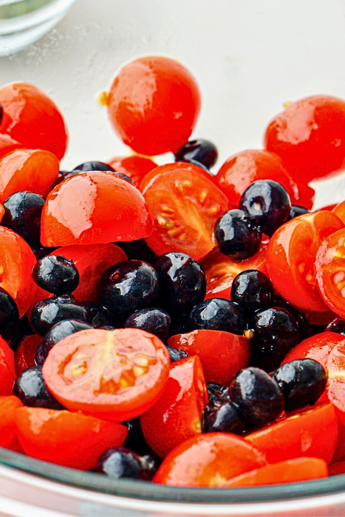 Blueberries and cherry tomatoes in a clear glass bowl tossed with oil and balsamic vinegar.