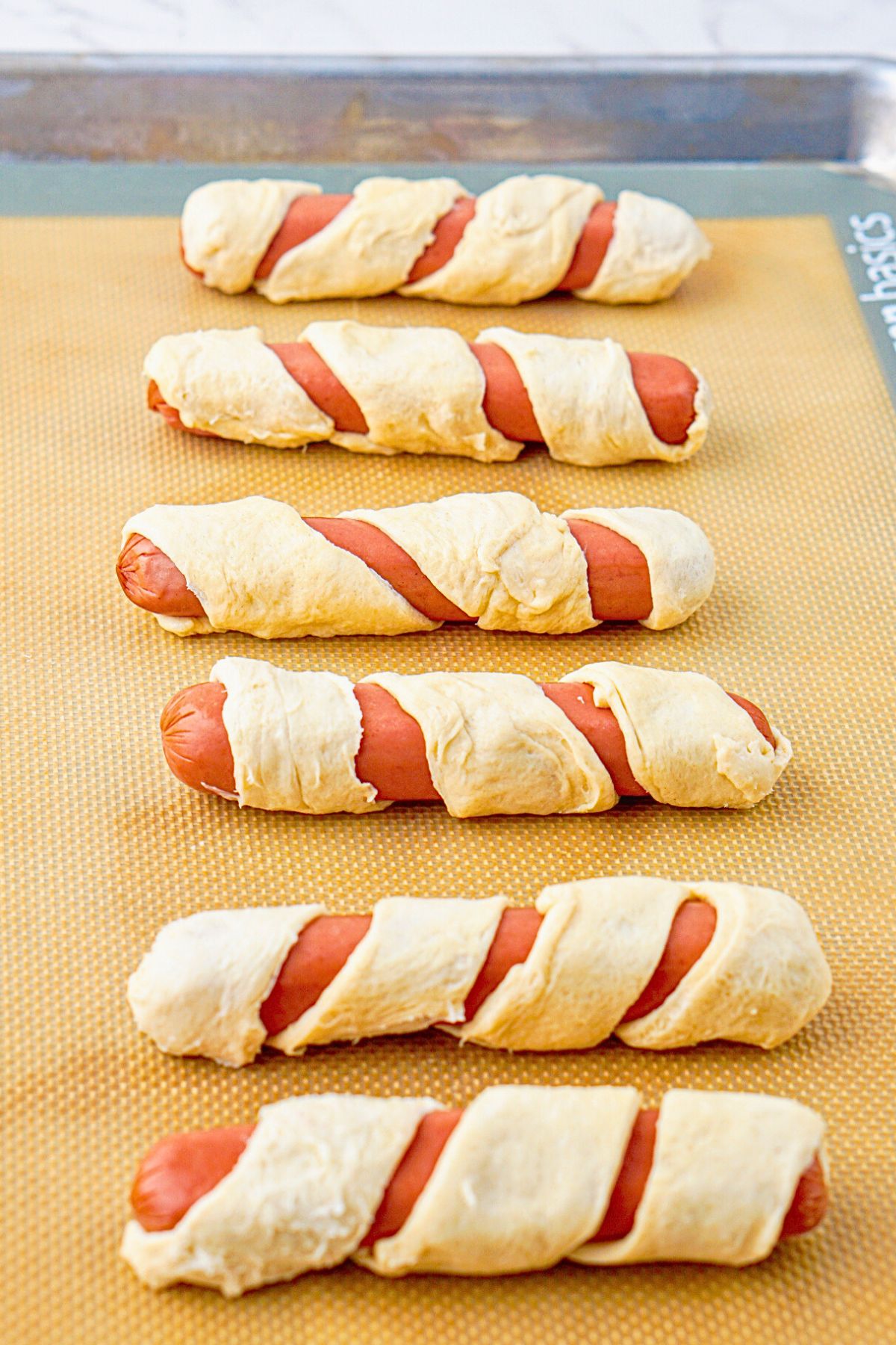 Hot dogs wrapped in strips of crescent roll dough before baking. 