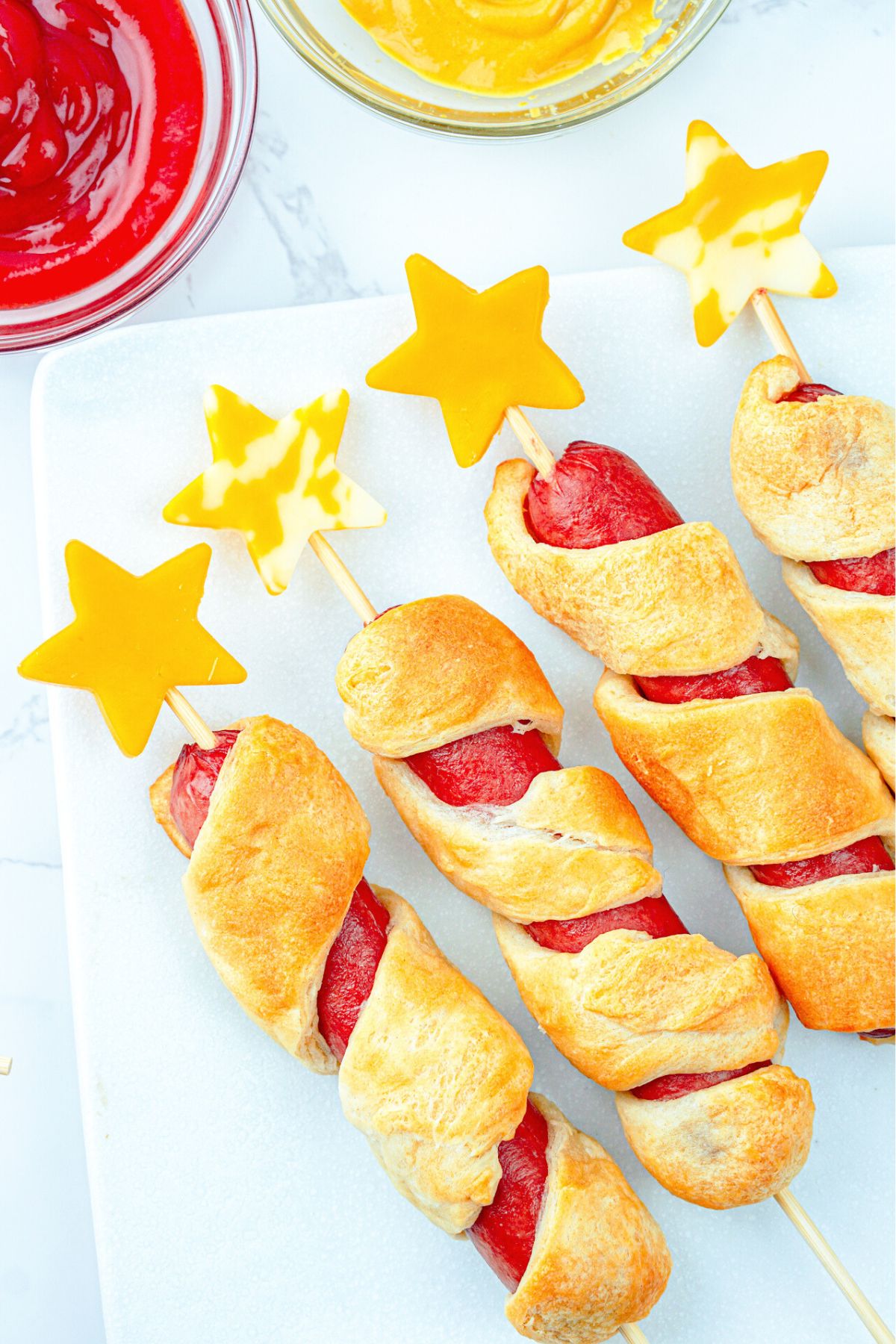 Dough wrapped hot dogs with cheese stars stuck into skewers, served with ketchup and mustard.