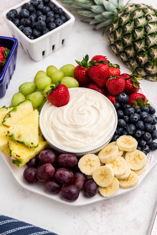 Bright berries, grapes, bananas, and pineapple, around a small bowl with fruit dip in the center of a large white platter.