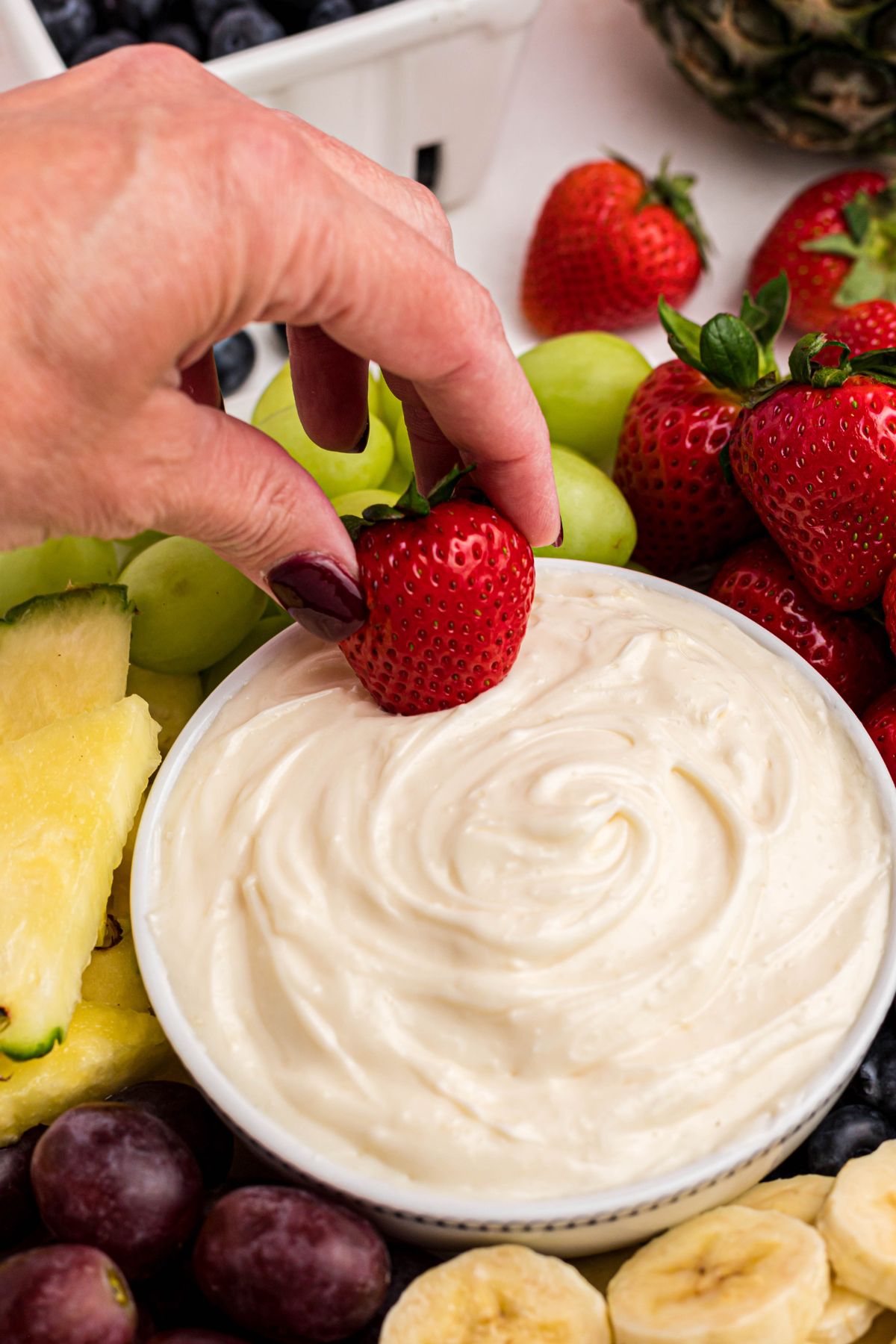 A hand dipping a bright red strawberry in a bowl of white creamy dip, surrounded by fruits. 
