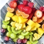 Pieces of fruit skewered in a rainbow color pattern on a small light gray cutting board.