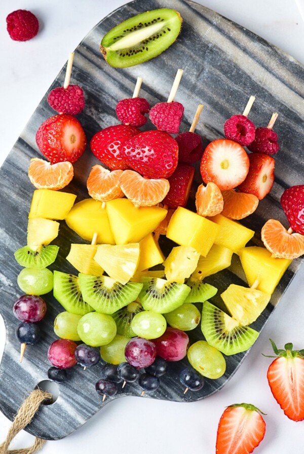 Pieces of fruit skewered in a rainbow color pattern on a small light gray cutting board.