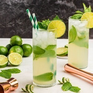 Bright yellow drink in a tall chilled glass, with two green striped straws, with limes and lime curls on the marble table.