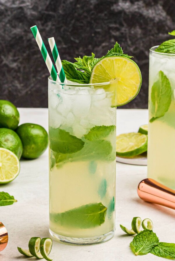 Bright yellow drink in a tall chilled glass, with two green striped straws, with limes and lime curls on the marble table.