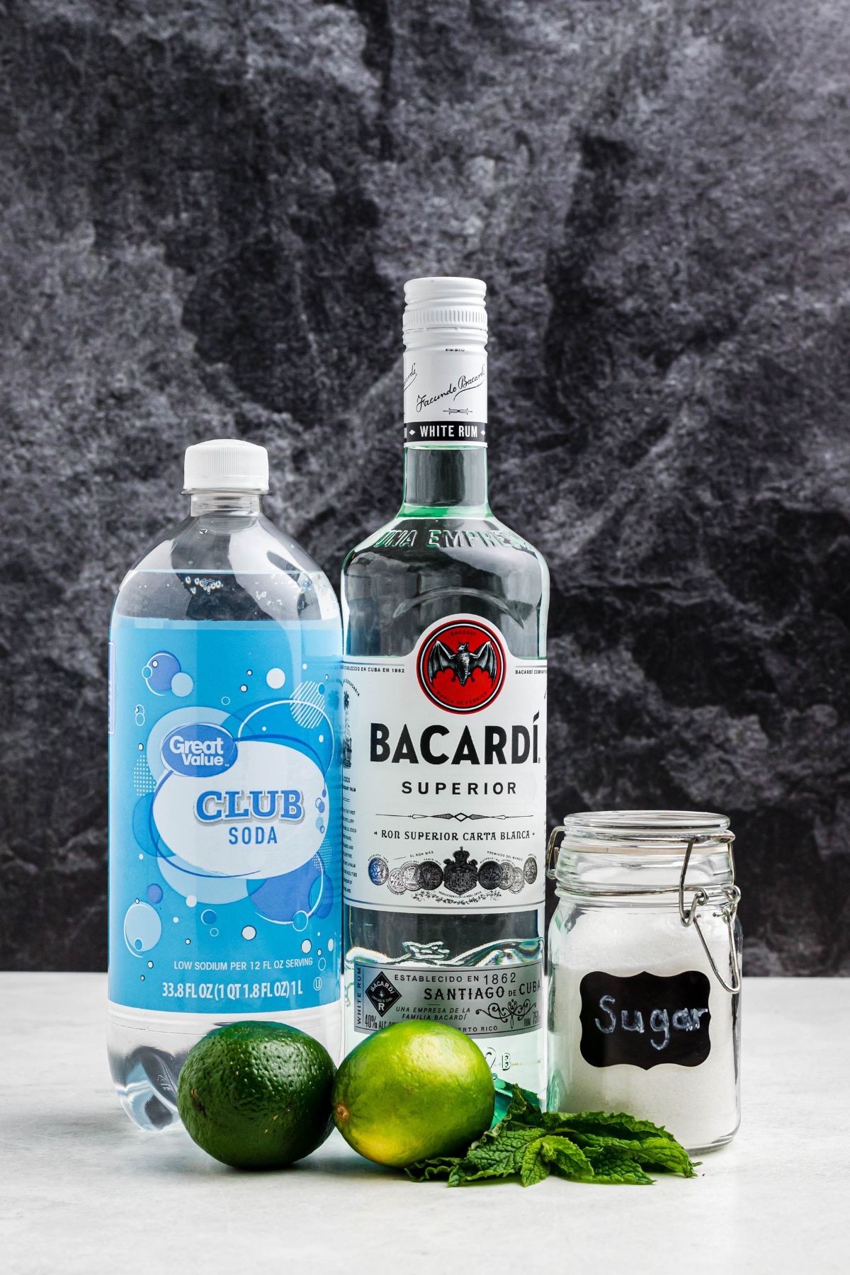 Bottles of rum and sparkling soda, with a jar of sugar, limes, and mint leaves, on a marble table. 