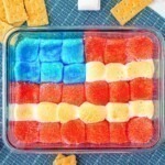 Red white and blue marshmallows over chocolate in a clear baking dish with graham crackers on the table for dipping.