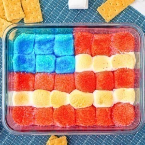 Red white and blue marshmallows over chocolate in a clear baking dish with graham crackers on the table for dipping.