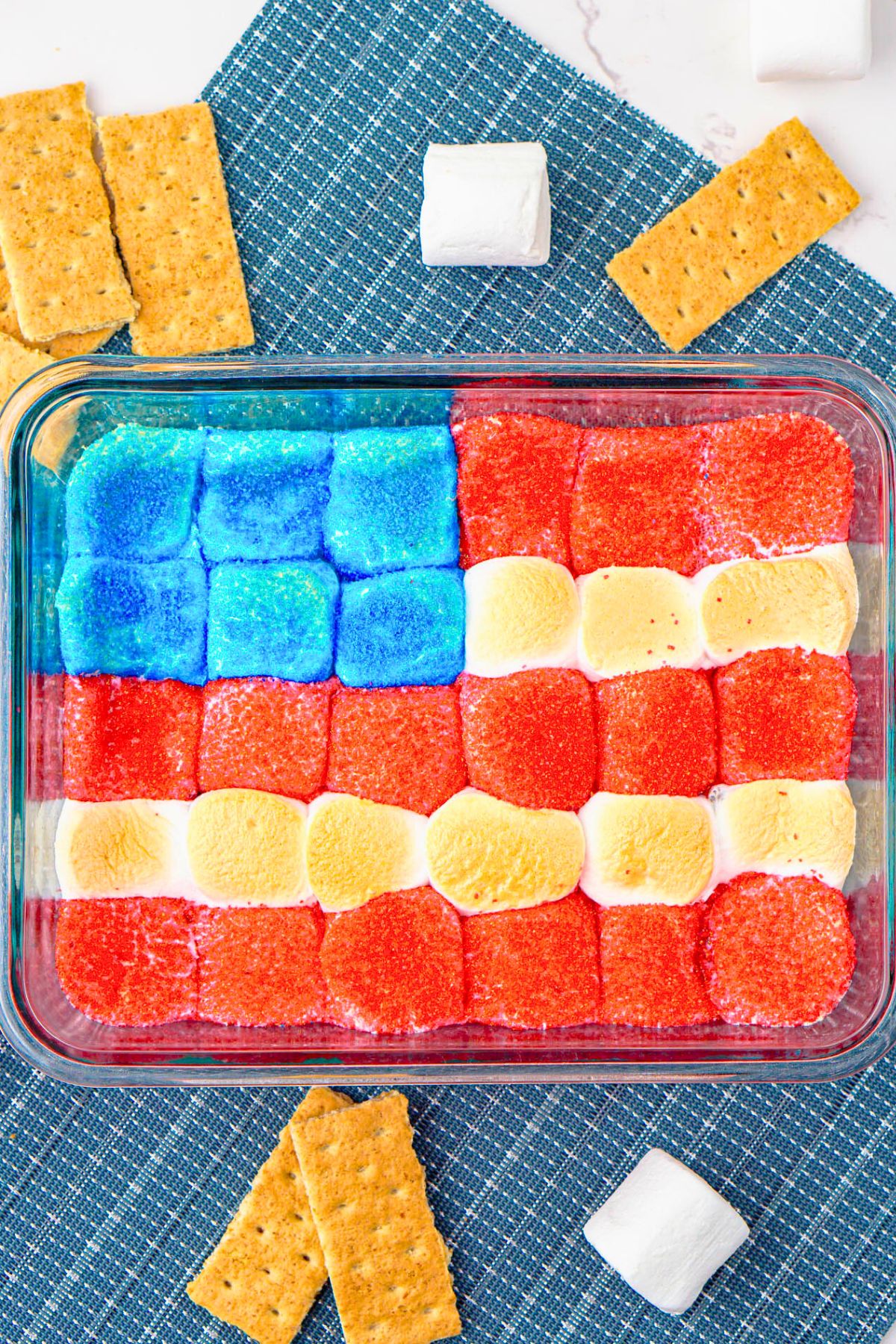 Red white and blue marshmallows over chocolate in a clear baking dish with graham crackers on the table for dipping.