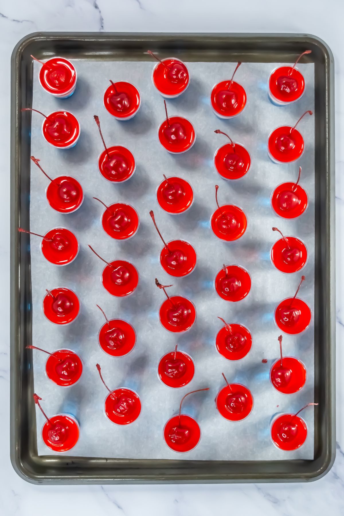 Jello cups topped with layers of jello and vodka mixture with red on top, on a baking sheet. 