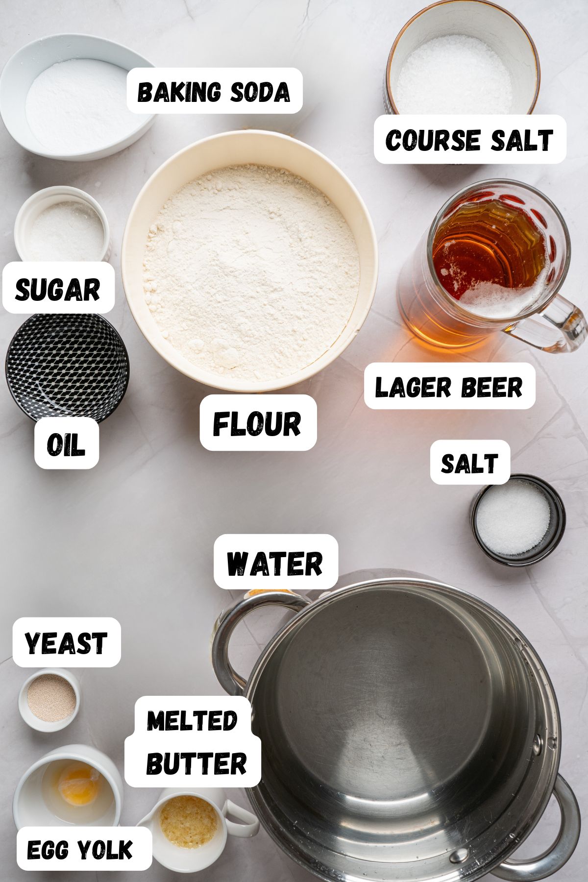 Soft pretzel ingredients like yeast, salt, and water, measured into small bowl on a white marble table, with labels of each ingredient printed over them. 