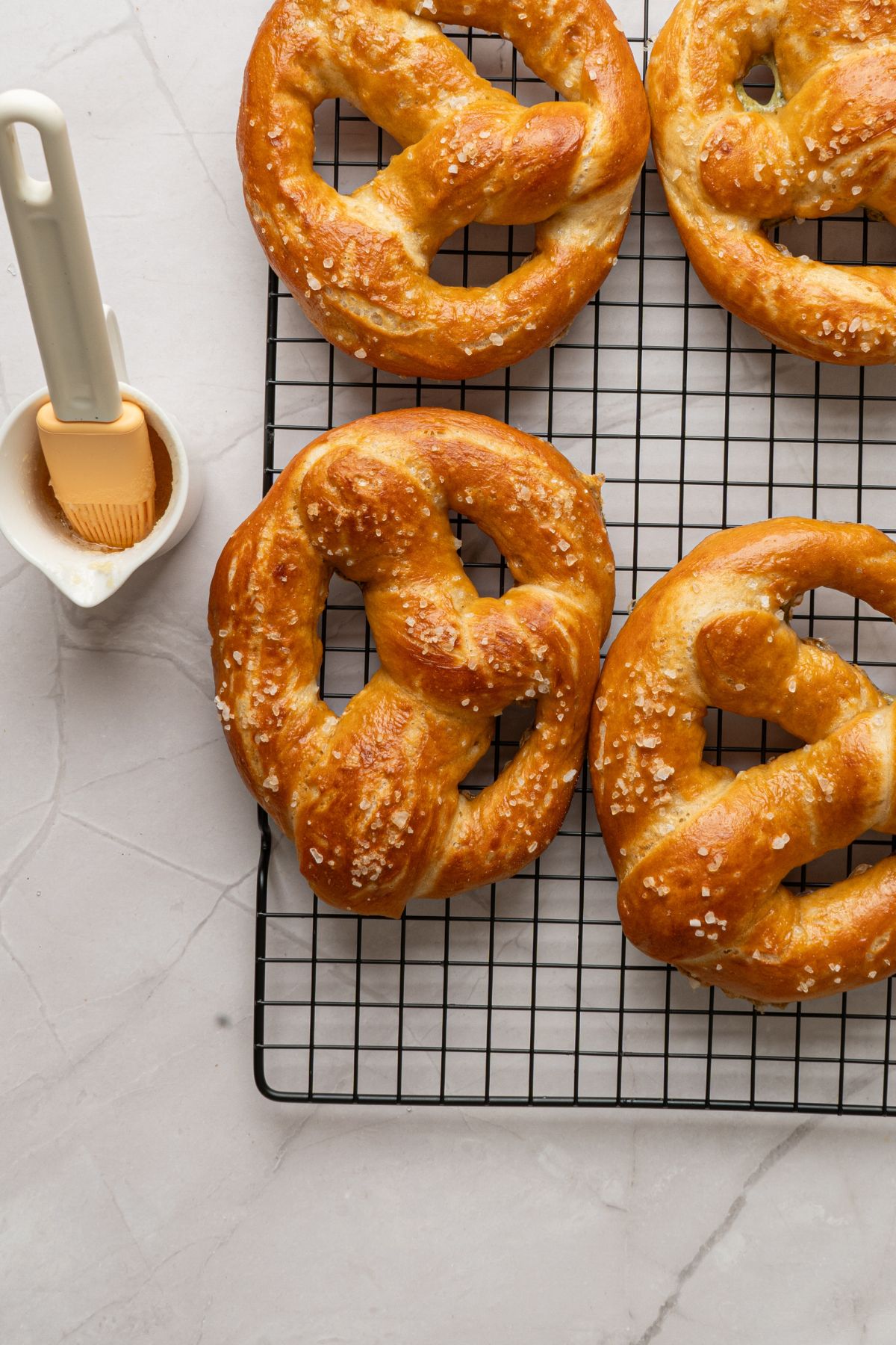 Baked golden pretzels on a wire rack on a marble table being brushed with butter. 