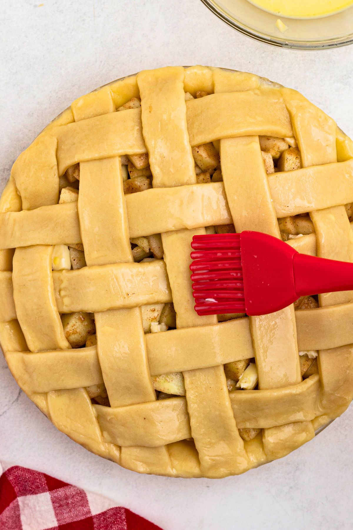 Uncooked apple pie with egg wash being brushed on top with a red pastry brush. 