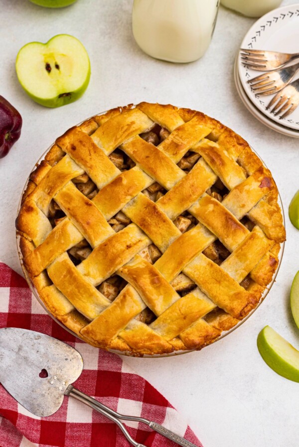 Golden crust, baked apple pie, on a white marble table, with slices of green apple, and a small plate with forks, next the whole pie.