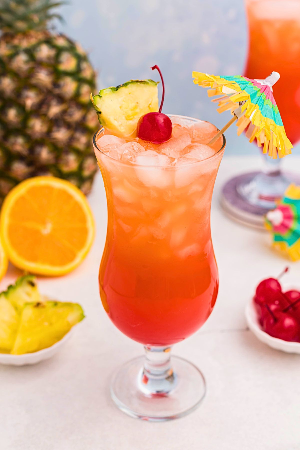 Red and orange drink mixed together, with a paper umbrella, in a hurricane glass with orange slices and cherries on the marble table.