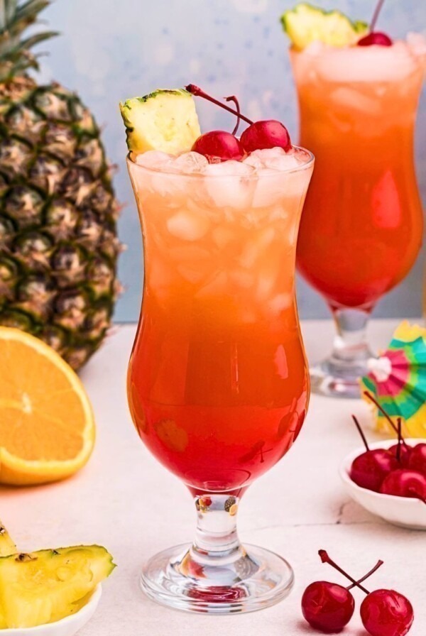 Red and orange drink mixed together in a hurricane glass with orange slices and cherries on the marble table.