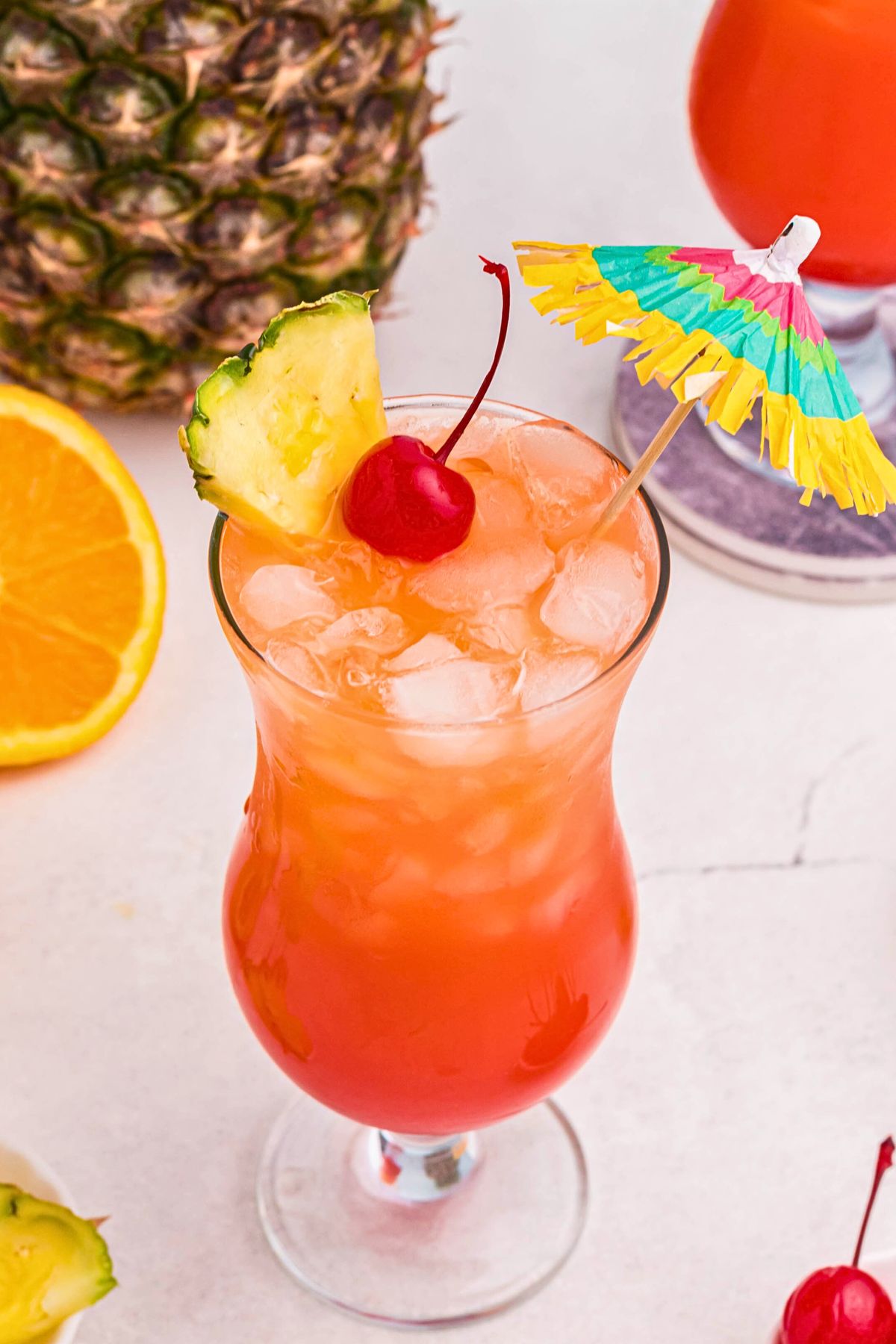 Red and orange drink in a hurricane glass, topped with a cherry, pineapple slice, a nd paper umbrella. 
