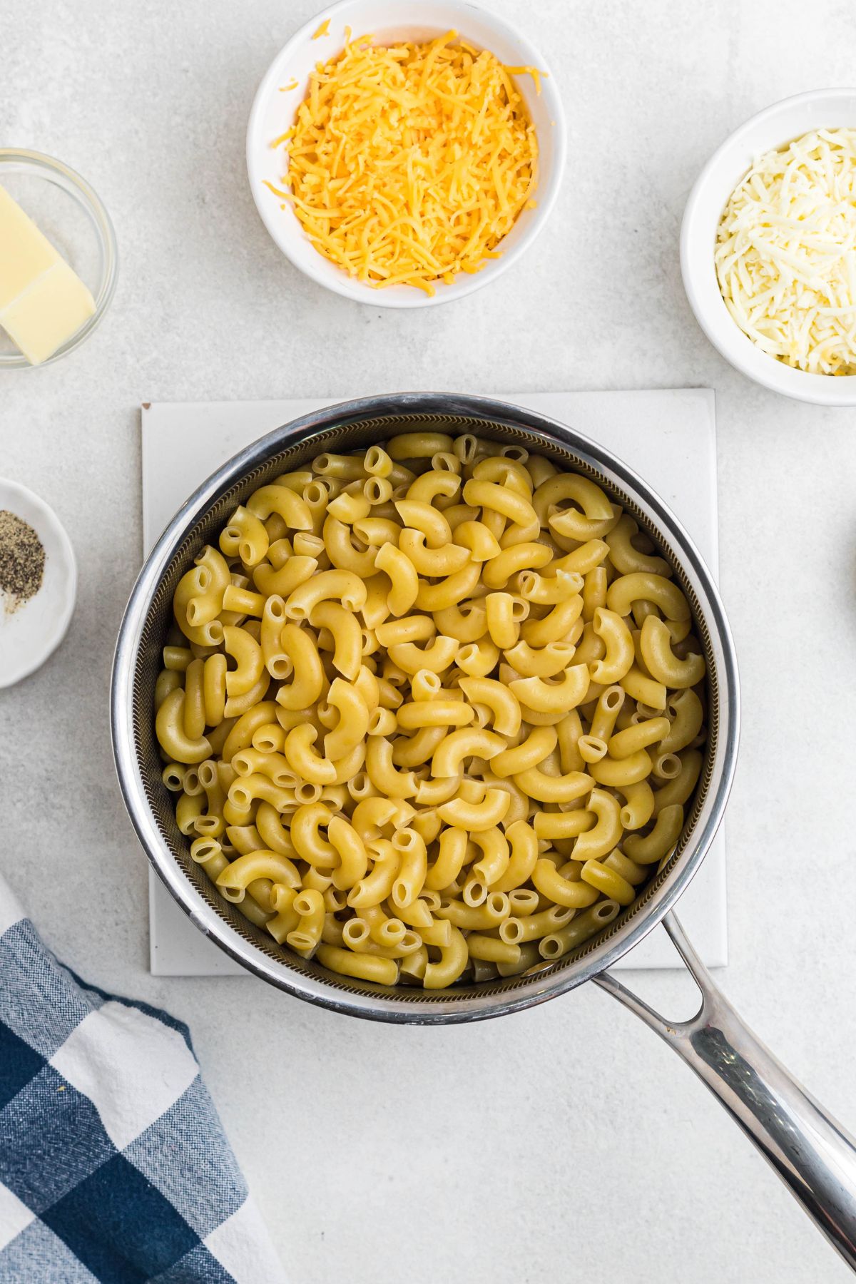 Lightly boiled elbow macaroni in a saucepan, surrounded by shredded cheese and seasonings.