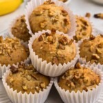 Light golden brown banana bread muffins in white liners, topped with chopped pecans.