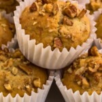 Light golden brown banana bread muffins in white liners, topped with chopped pecans.
