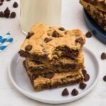 Cookie bars stacked on a small white plate in front of a tall glass of milk, with chocolate chips scattered on the table.
