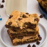 Cookie bars stacked on a small white plate in front of a tall glass of milk, with chocolate chips scattered on the table.