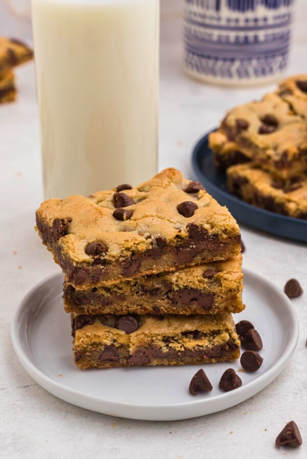 Golden chocolate chip cookie bars stacked on a small white plate with a tall glass of milk behind the plate.