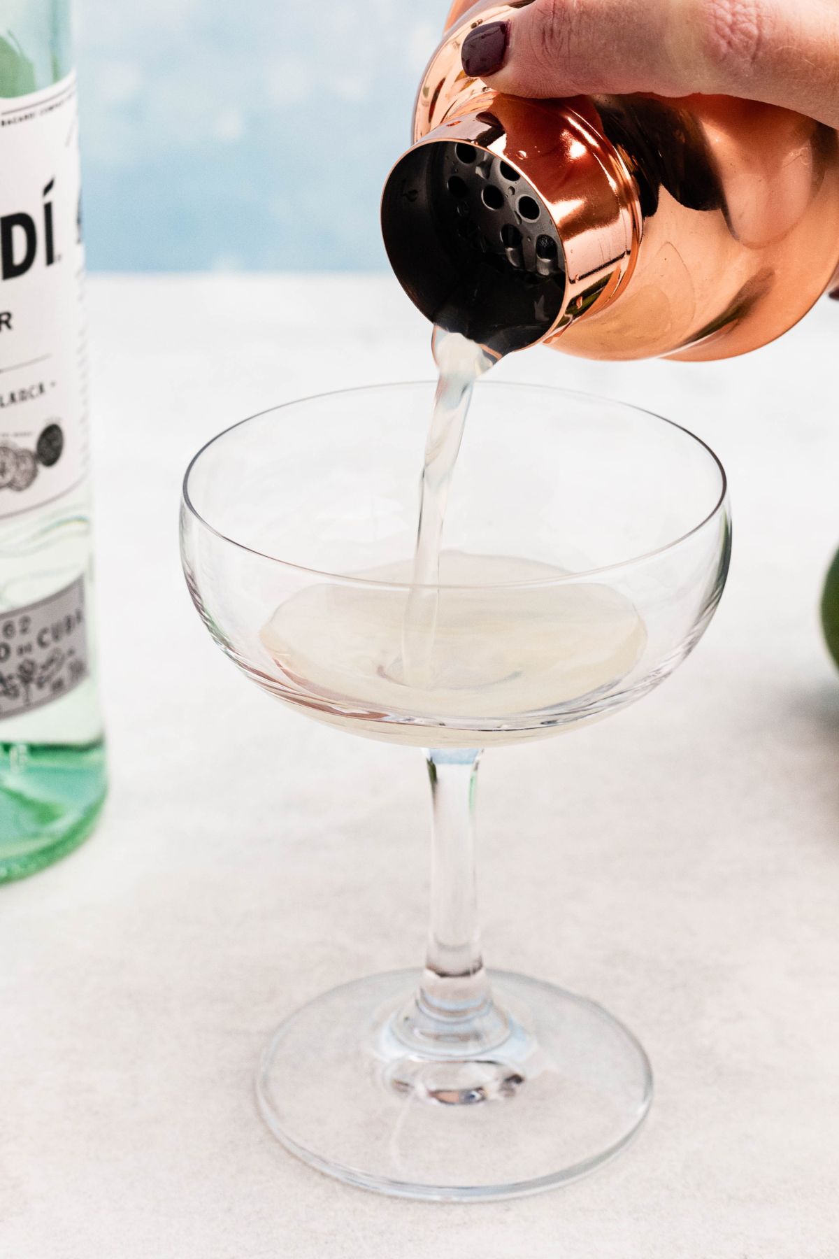 Mixed ingredients being poured through a strainer on a cocktail shaker into a small coupe glass. 