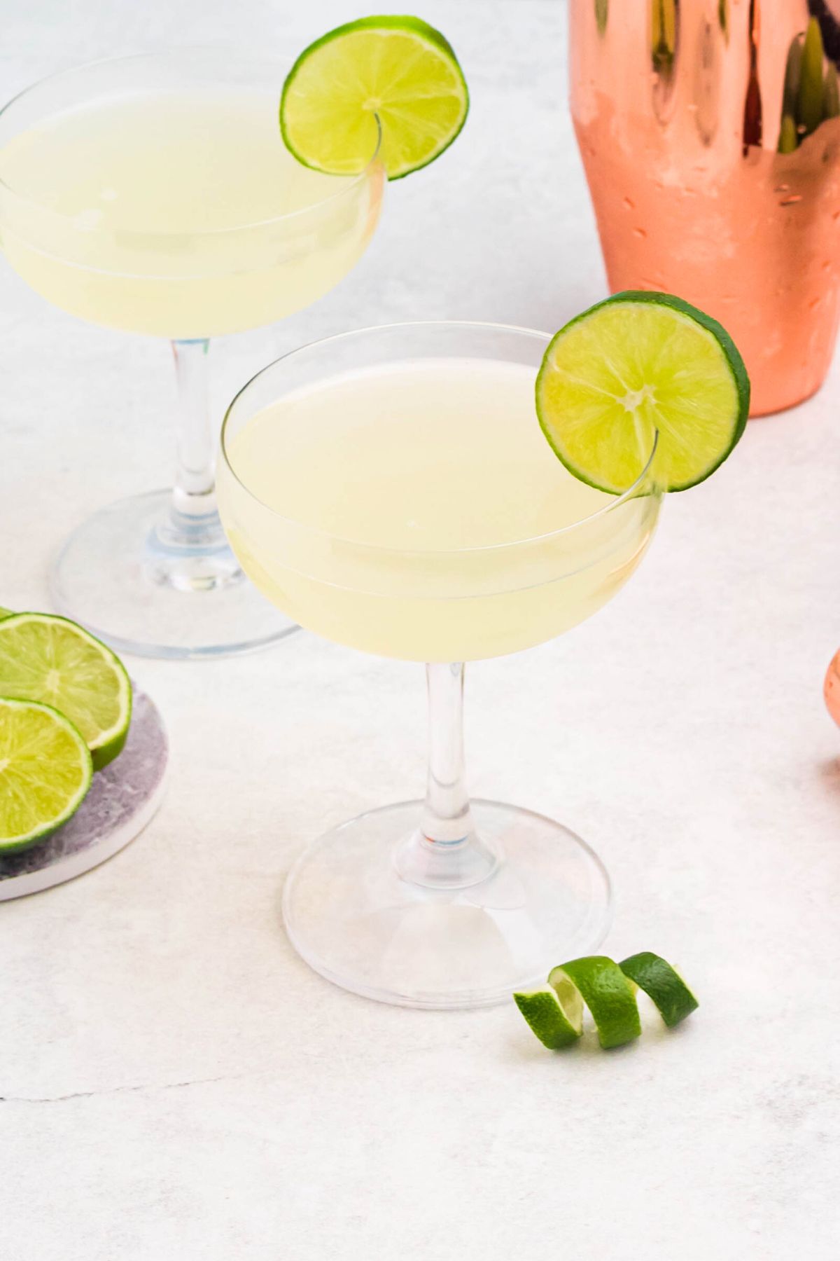 Small glass filled with drink, garnished with lime wheel, on a marble table with limes and lime curls on the table. 