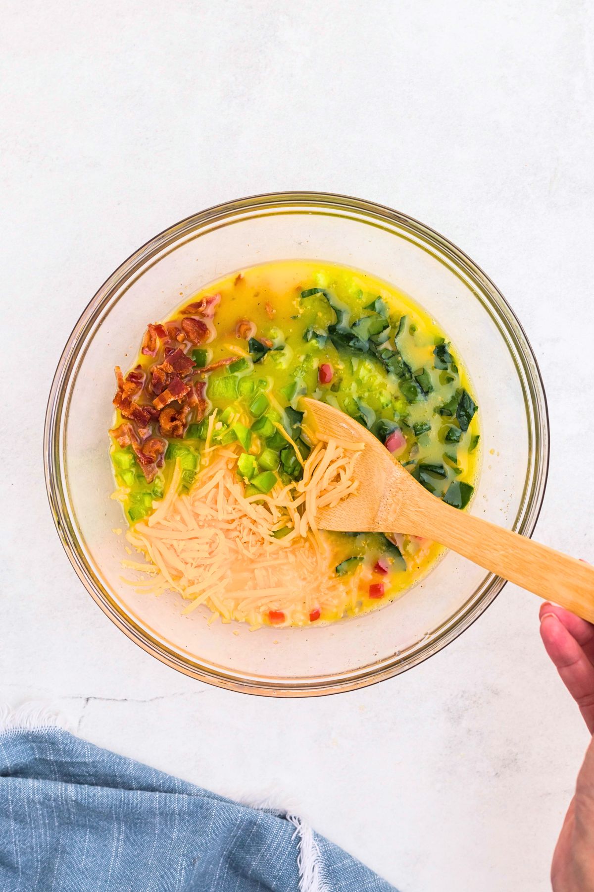 Bell peppers, cheese, and other ingredients being mixed with whisked eggs in clear glass bowl. 