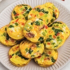 Bright yellow egg bites with red and green chopped bell peppers baked inside, on a round cream plate.