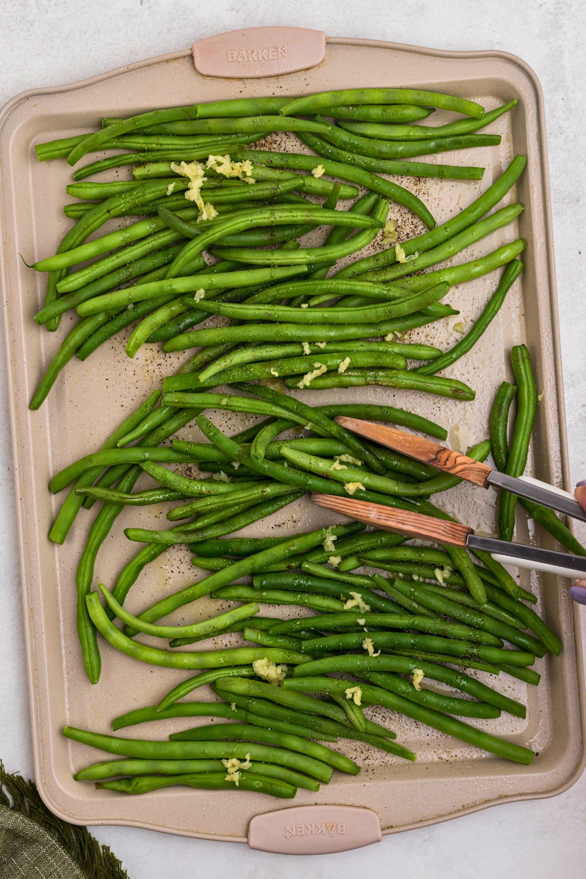 Fresh green beans on a baking sheet, being tossed with tongs with olive oil and seasonings. 