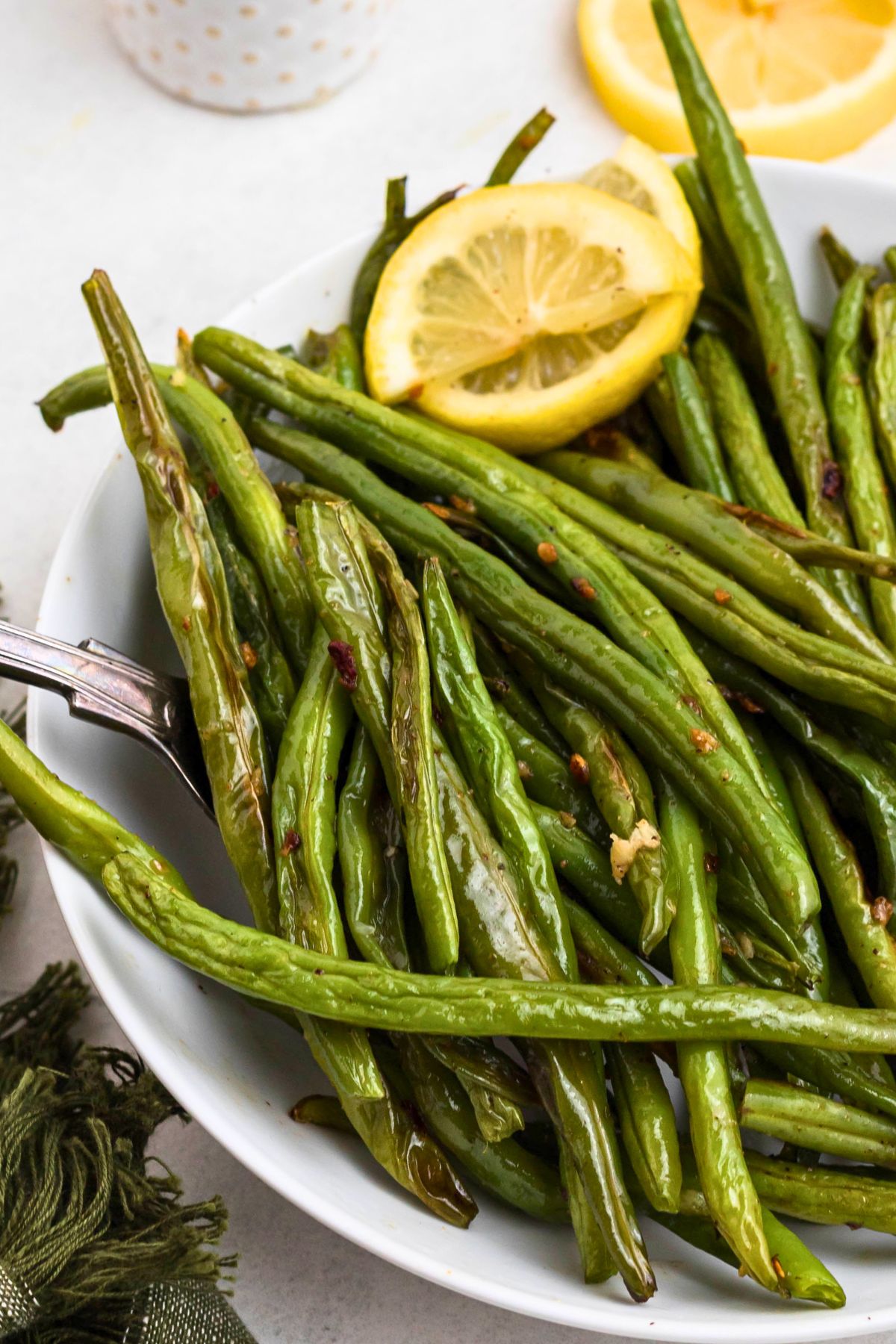 Fresh green beans in a white bowl, with a spoon lifting a serving  of beans and fresh lemons on the side. 