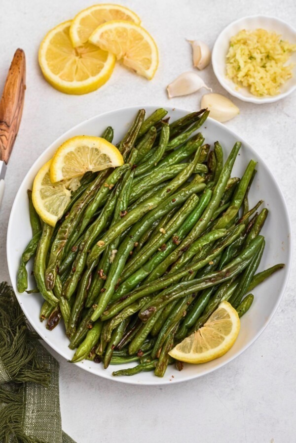 Bright green roasted green beans, in a white bowl, with lemon wedges as a garnish.