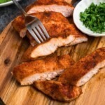 Smoked, seasoned chicken on a small, round cutting board, with parsley flakes in a small cutting board.