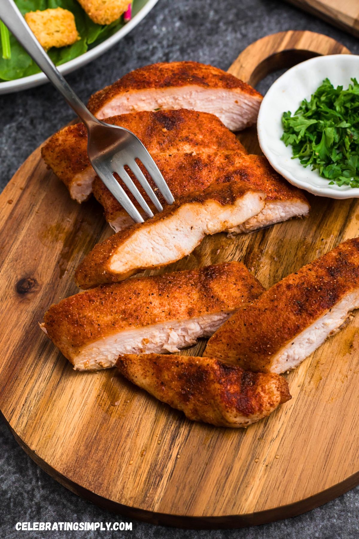 Smoked, seasoned chicken on a small, round cutting board, with parsley flakes in a small white bowl. 