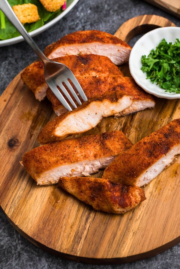 Smoked, seasoned chicken on a small, round cutting board, with parsley flakes in a small white bowl.