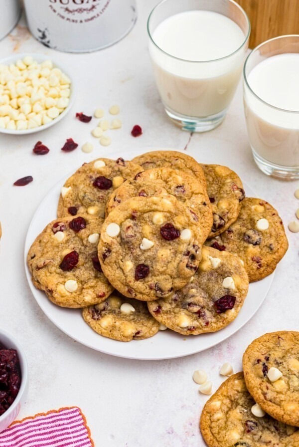 White chocolate chip and cranberry cookies stacked on a white plate with glasses of milk on the table.