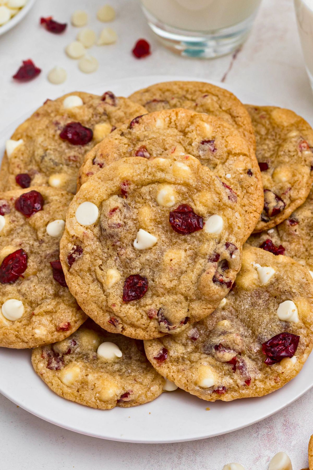 White chocolate chip and cranberry cookies stacked on a white plate with glasses of milk on the table.