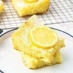 Bright yellow lemon bars stacked on a small white plate with a slice of lemon on top.